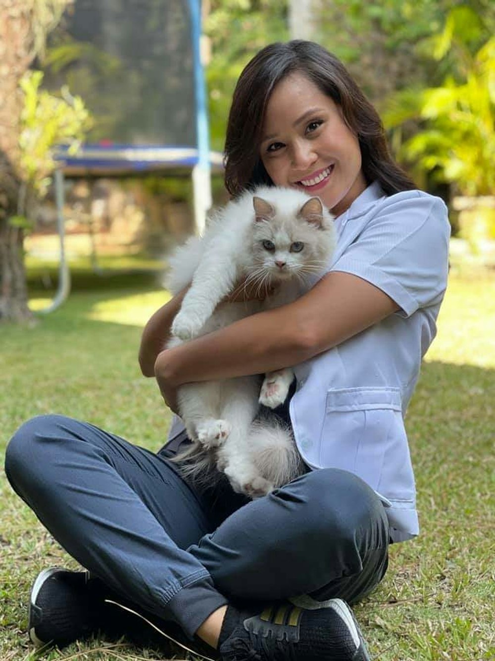 Veterinária sorridente sentada na grama abraçando um gato branco